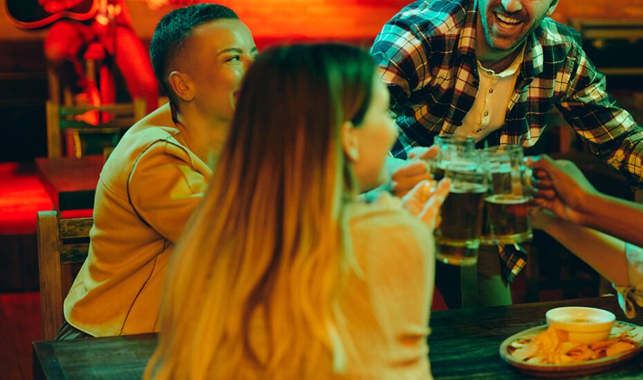 Young cheerful man and his friends having fun while drinking beer and toasting during the night out in a pub.